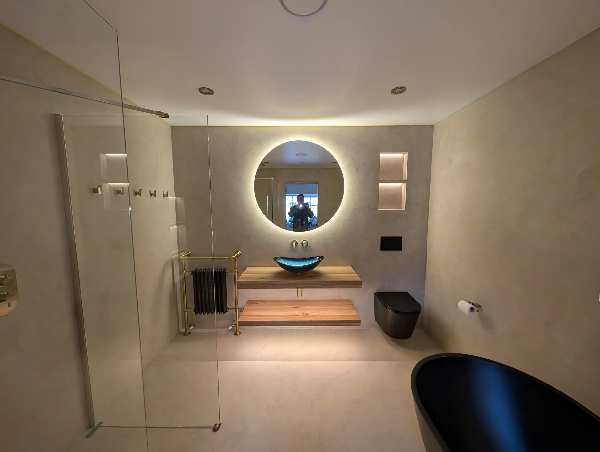 Completed microcement bathroom with an illuminated round mirror, floating oak vanity and walk-in shower